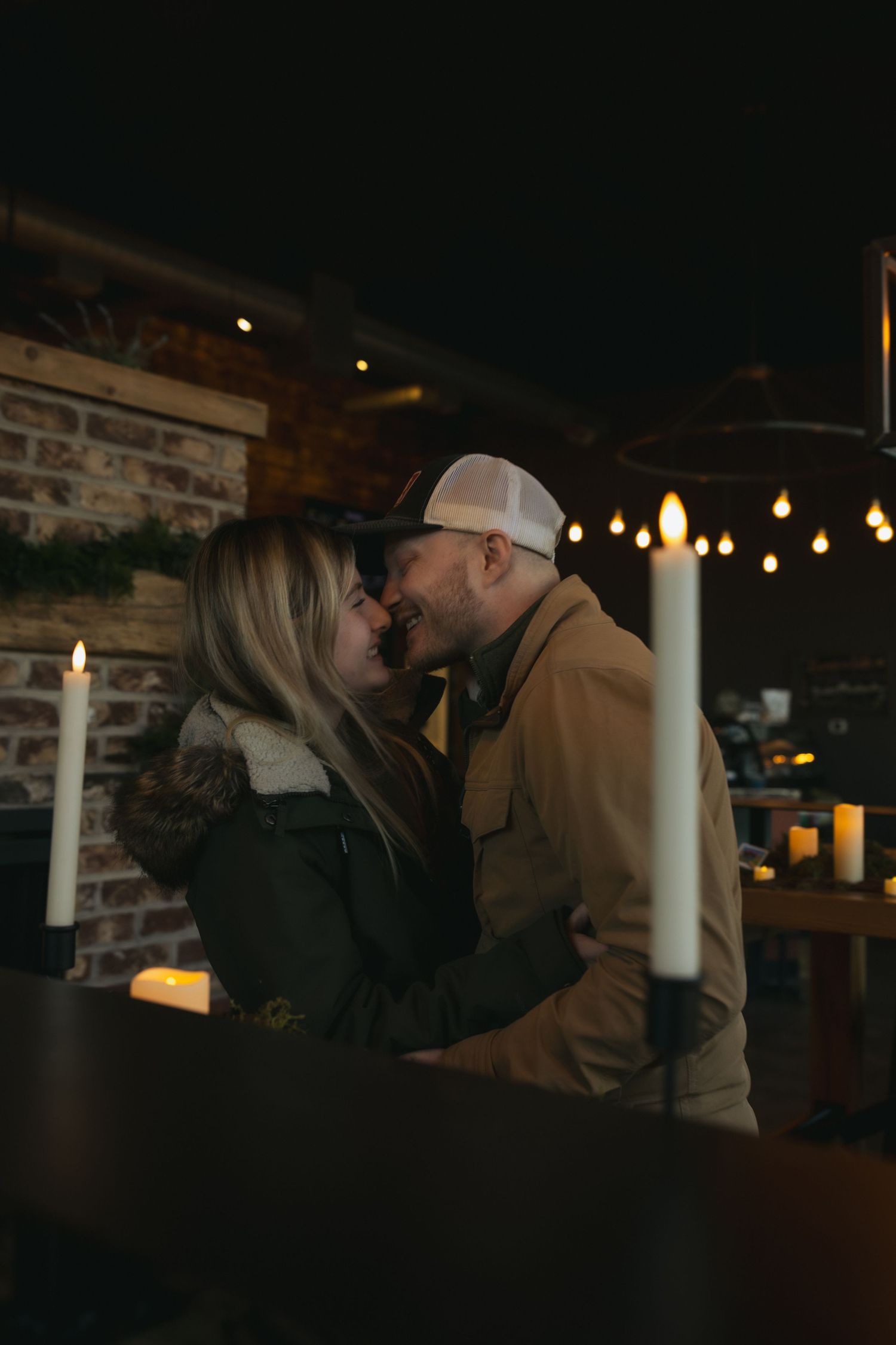 A romantic couple shares an intimate moment at a dimly lit restaurant with candles and string lights creating ambiance.