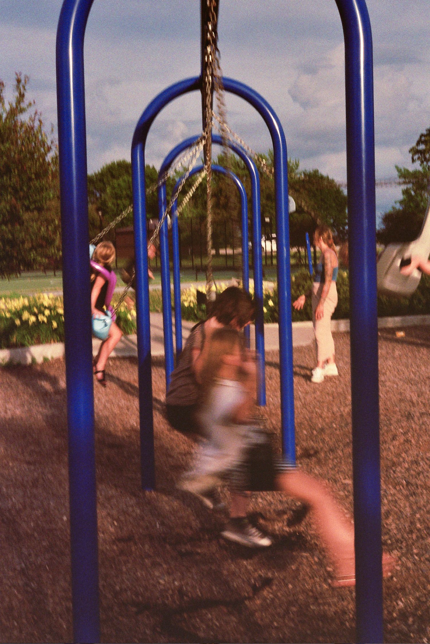 Children play on a row of blue metal swing set arches in a playground with flowers visible in the background.