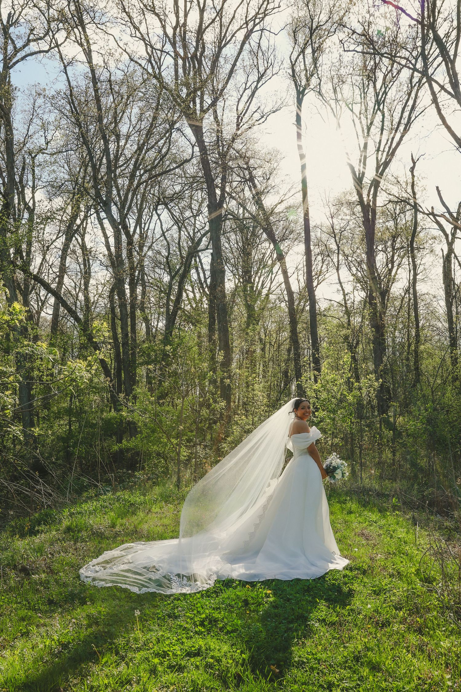 A wedding dress with a flowing veil creates an ethereal scene in a spring forest with budding green foliage.