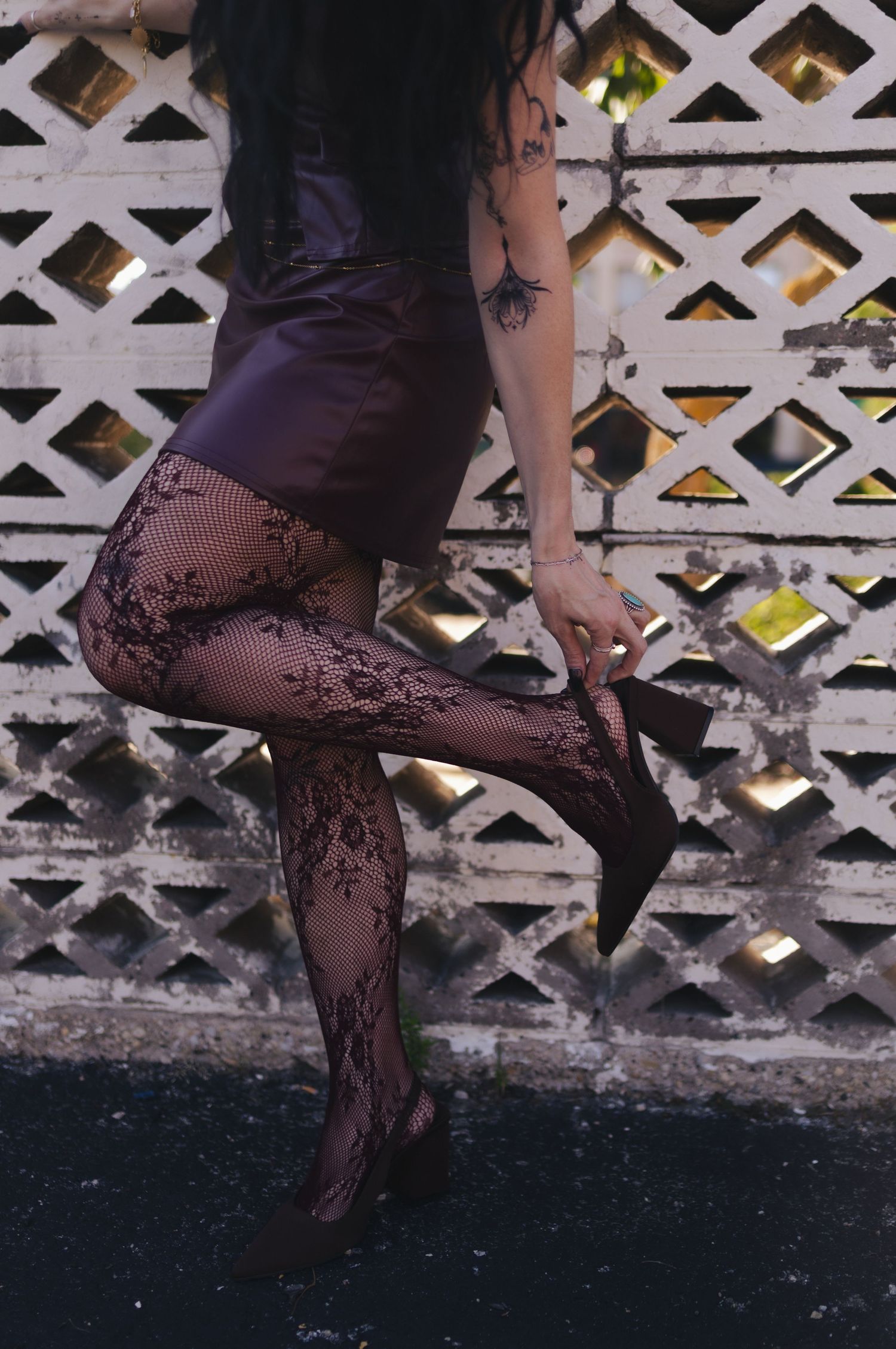 Woman in purple mini skirt and floral lace tights holding a black heel shoe near a white lattice fence.