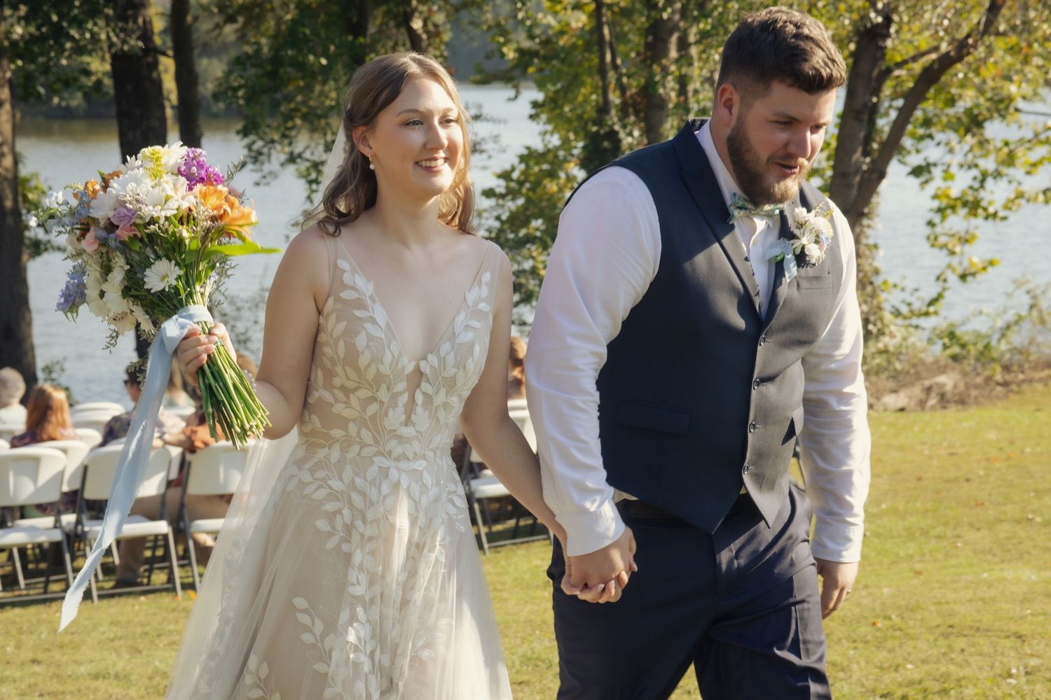 Newlyweds walking hand in hand during outdoor lakeside wedding ceremony.