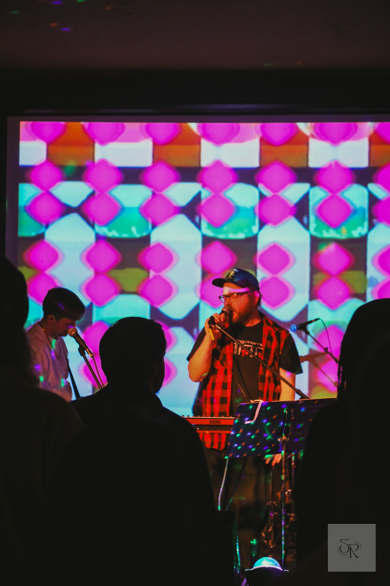 Performers on stage at a concert with vibrant pink and turquoise geometric patterns displayed on screen behind them.