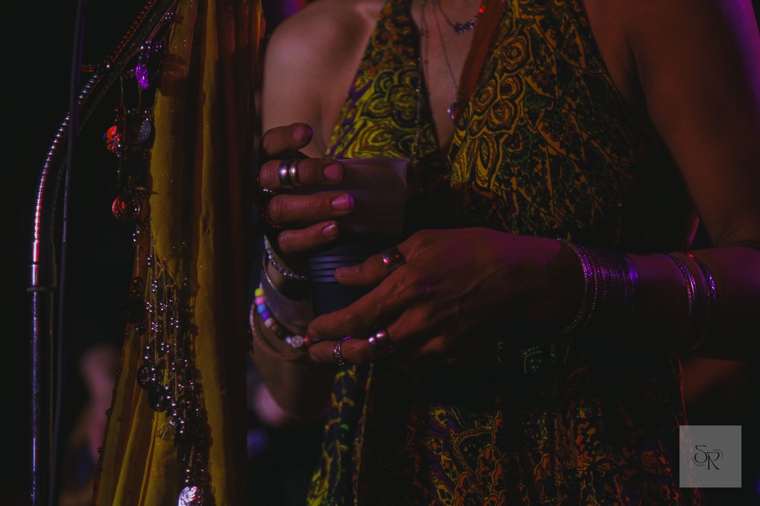 Someone holds a drink in a dark nightclub setting wearing a gold patterned dress and bangles.
