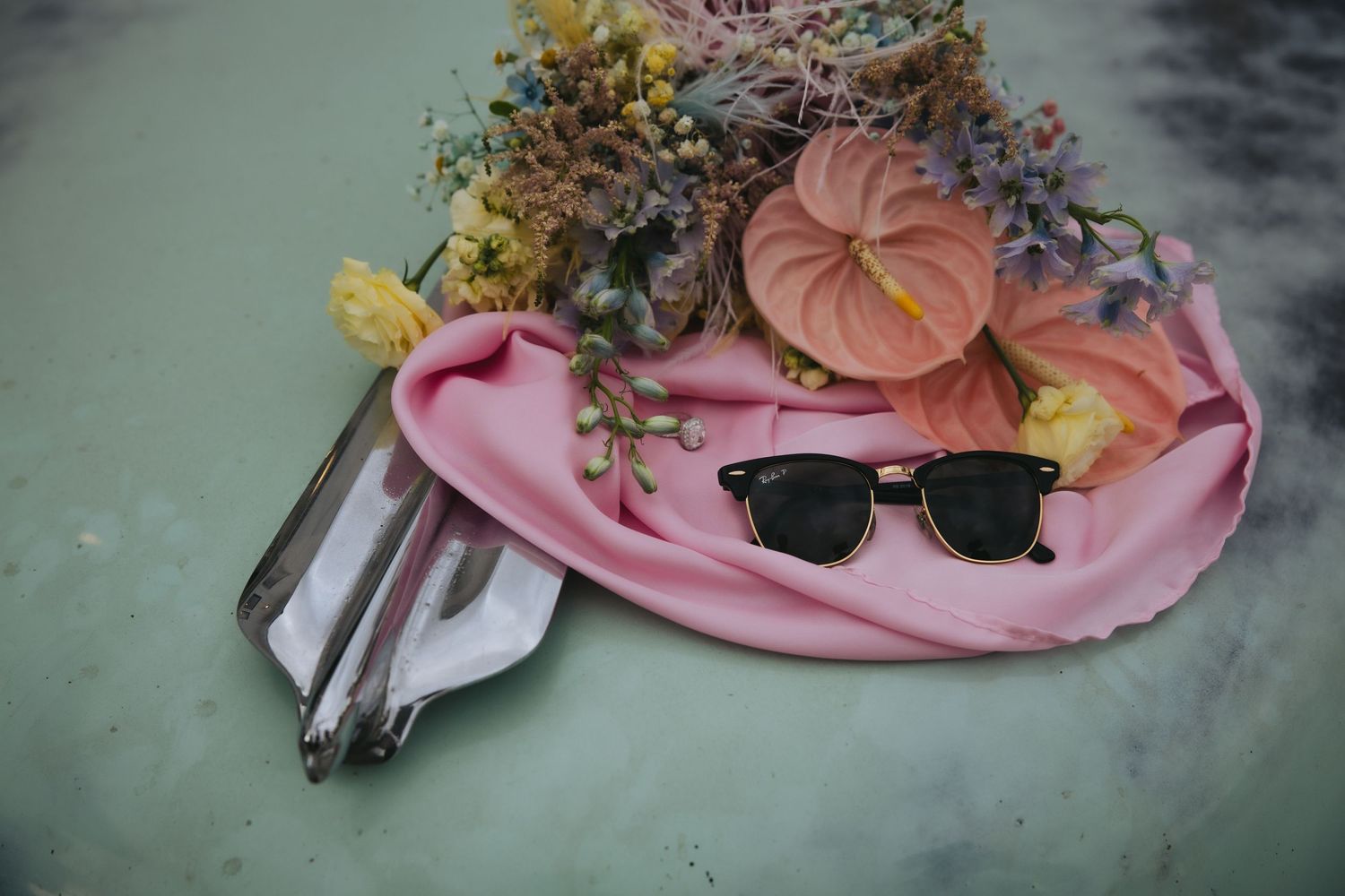 A pink silk scarf with sunglasses, decorative pumpkins and dried flowers arranged on a turquoise surface.