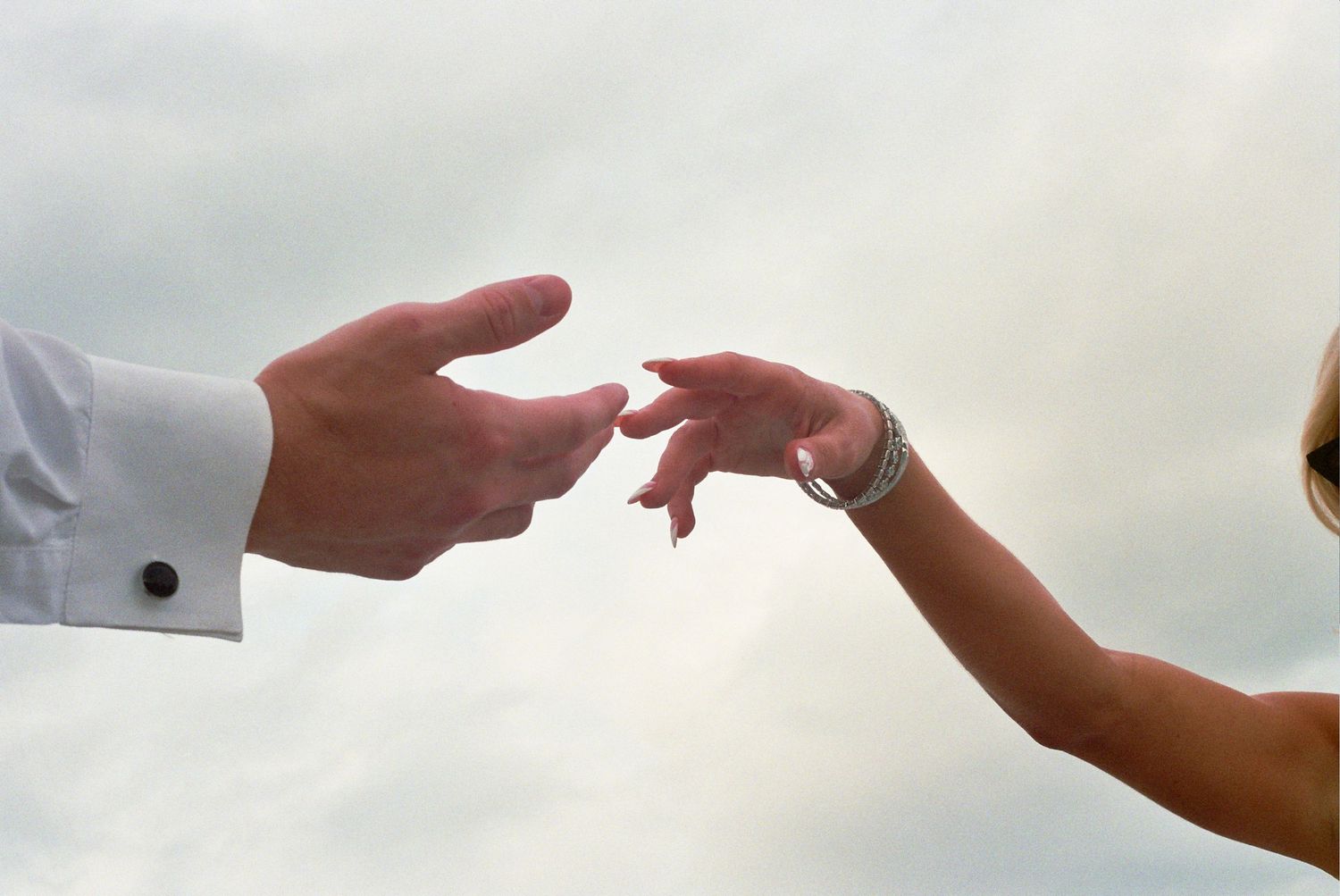 Two hands reach towards each other with fingertips nearly touching against a light cloudy sky background.