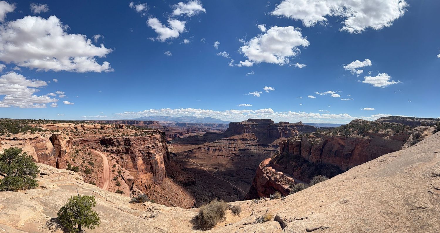 A panoramic view of a desert canyon landscape with red rock formations and white clouds in the blue sky above.