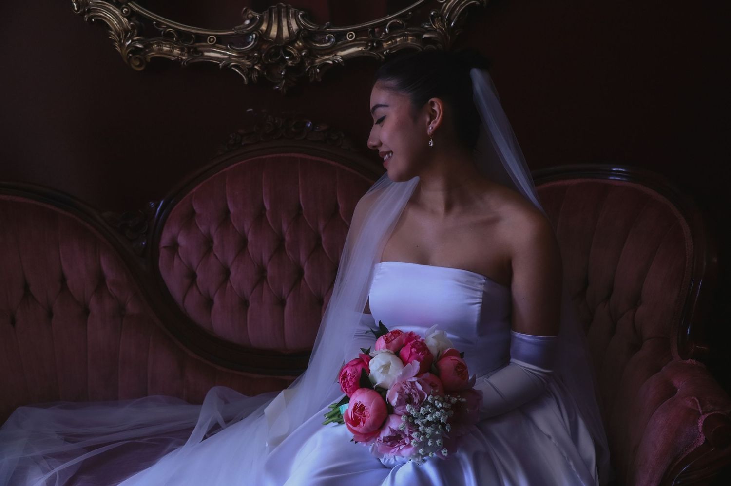 A bride in a white strapless gown sits elegantly on a burgundy tufted sofa holding a pink and coral floral bouquet.
