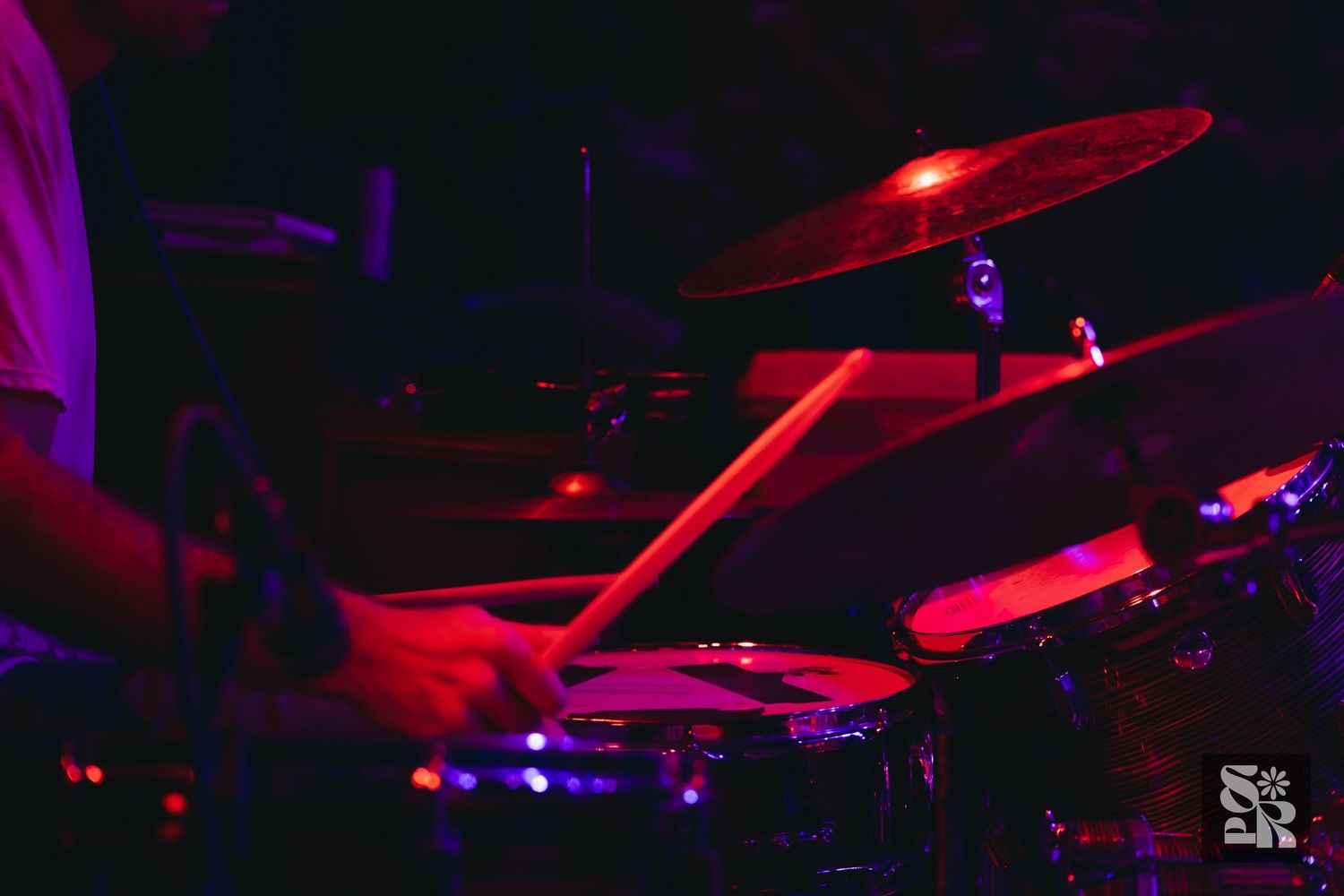 Drummer performing live on stage under red and purple lights, playing drum kit with cymbals visible.