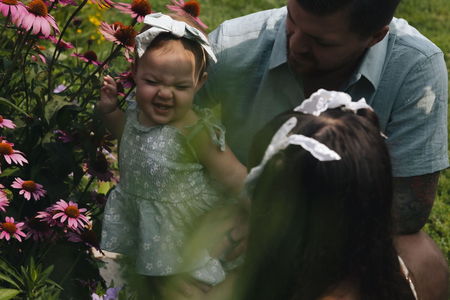 A joyful baby in a green dress held by parents among blooming pink coneflowers in a lush garden.
