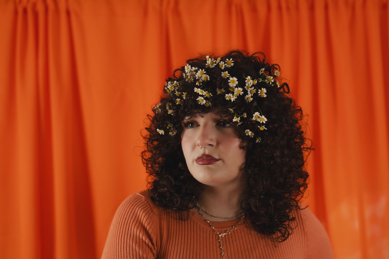Woman with curly black hair adorned with small white flowers, wearing an orange top against a bright orange background.