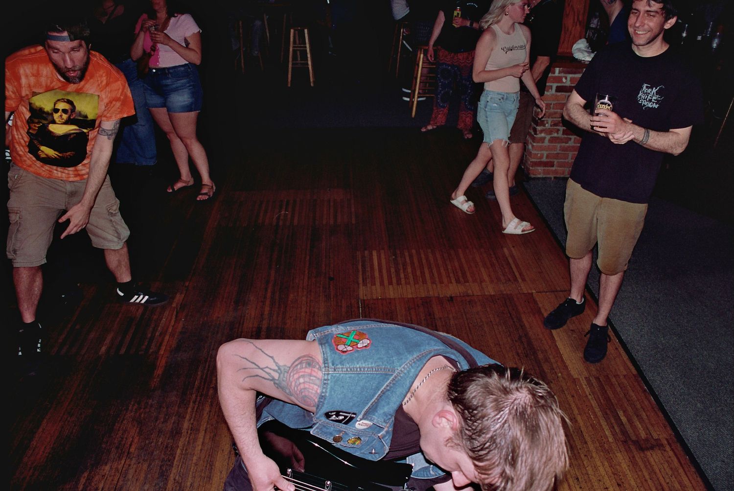 Person doing a dramatic floor move on a wooden dance floor surrounded by onlookers at a lively indoor party.