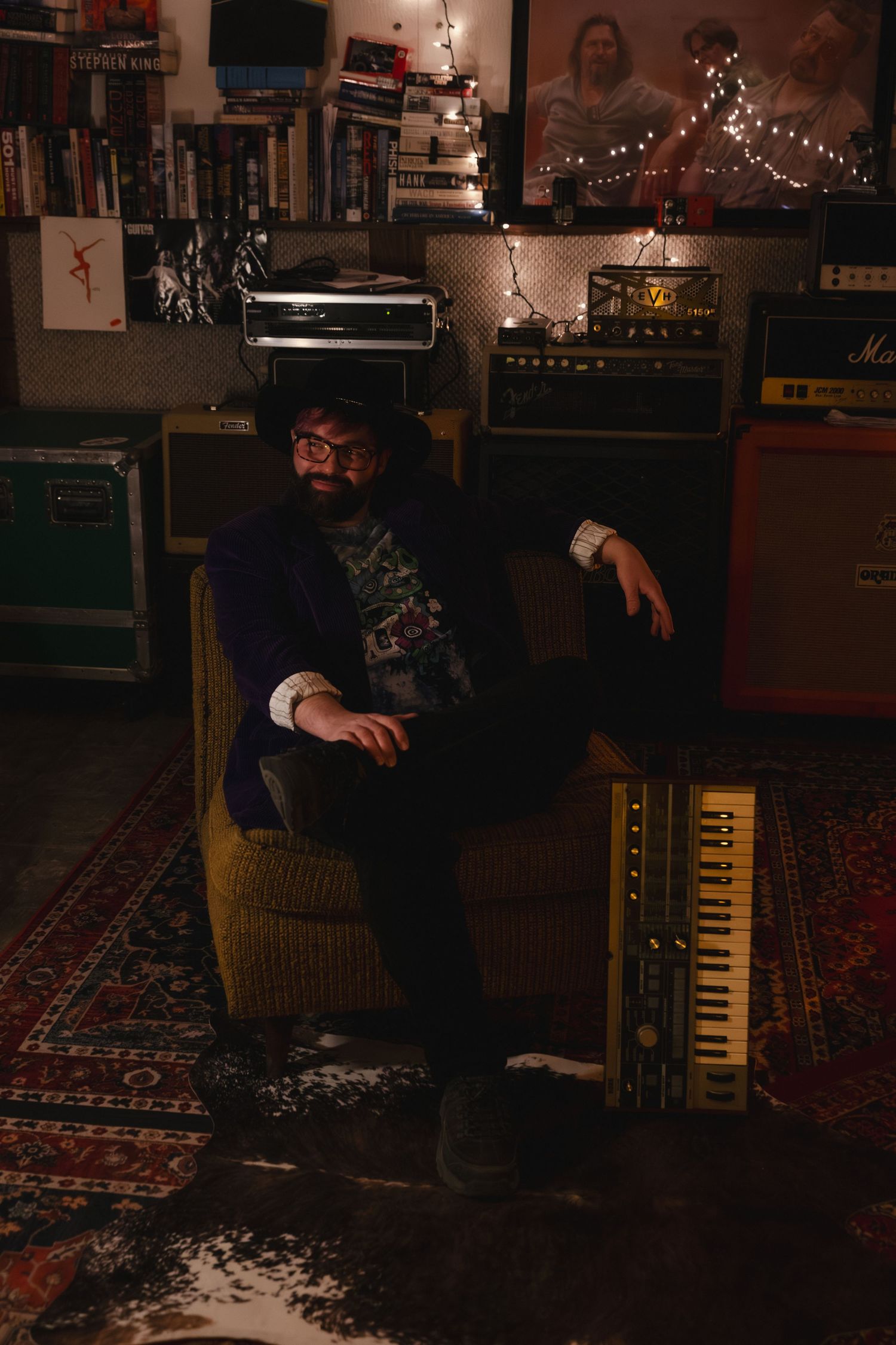 Bearded man in dark jacket sits in armchair beside a keyboard in a dimly lit music studio with amplifiers.