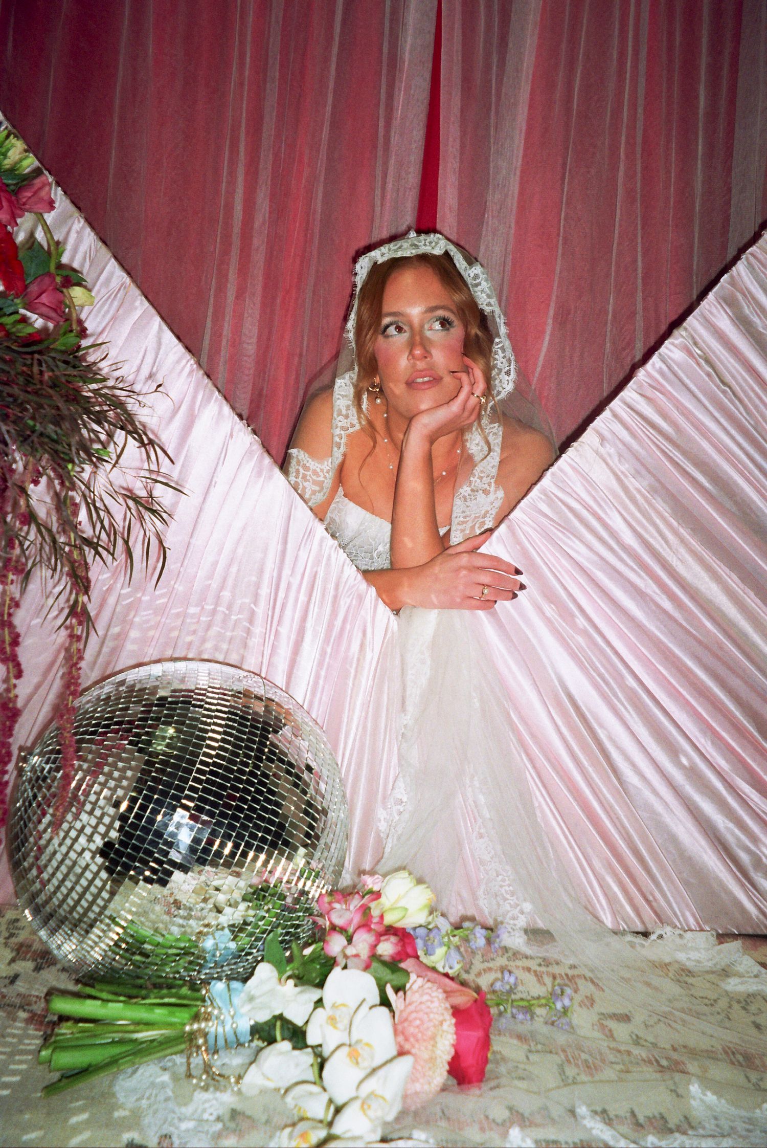 Bride in white lace gown and veil poses dreamily beside a disco ball and floral bouquet against pink draped curtains.