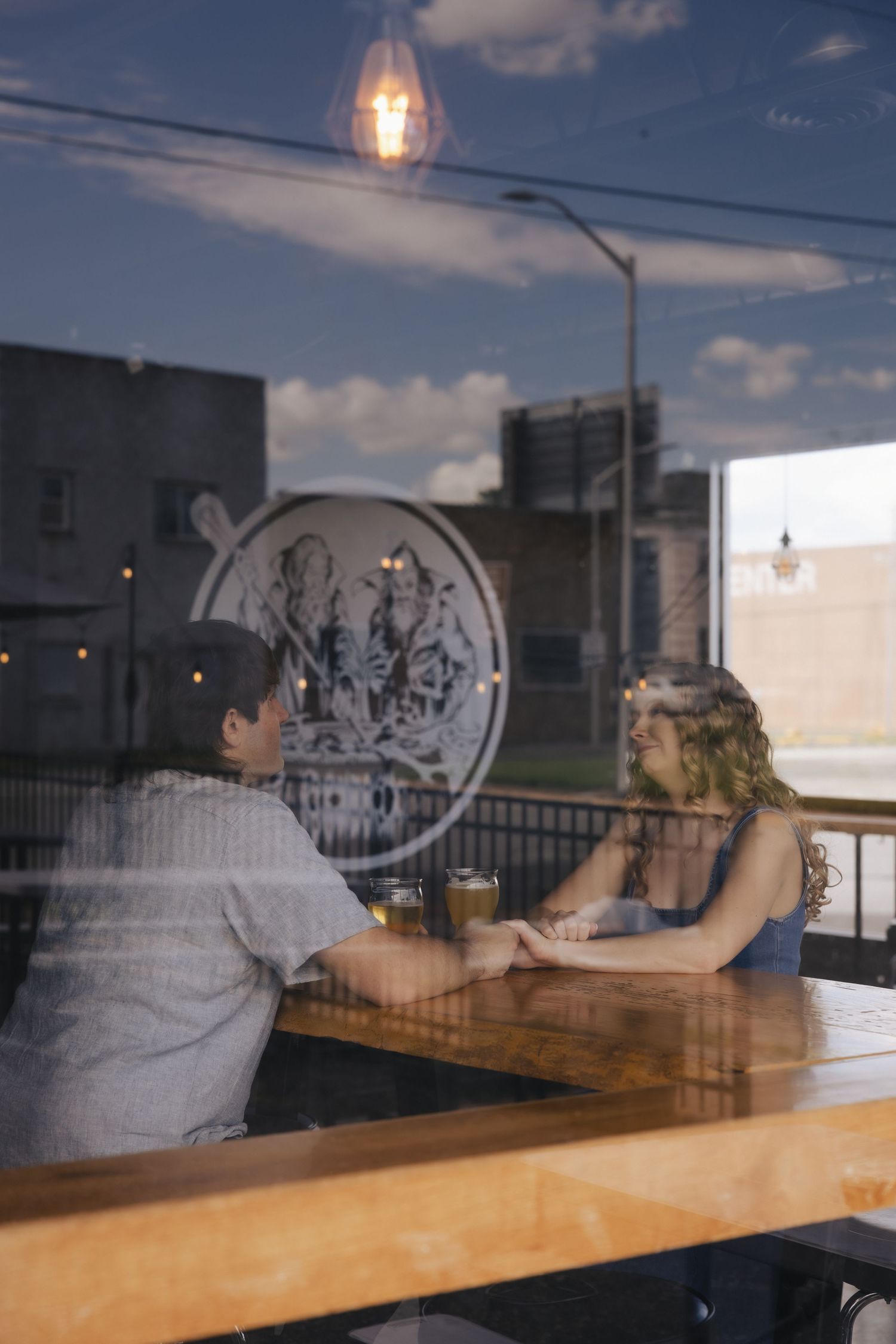 Couple holding hands at a wooden cafe table, viewed through a window reflecting city buildings and clouds.