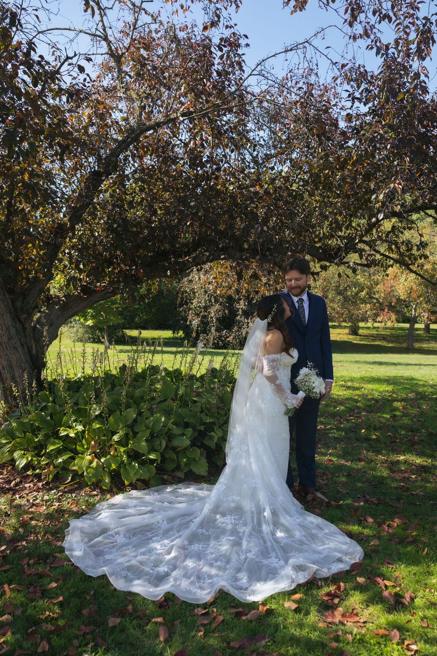 Elegant wedding couple poses in a scenic autumn orchard with fallen leaves and flowing white bridal gown.