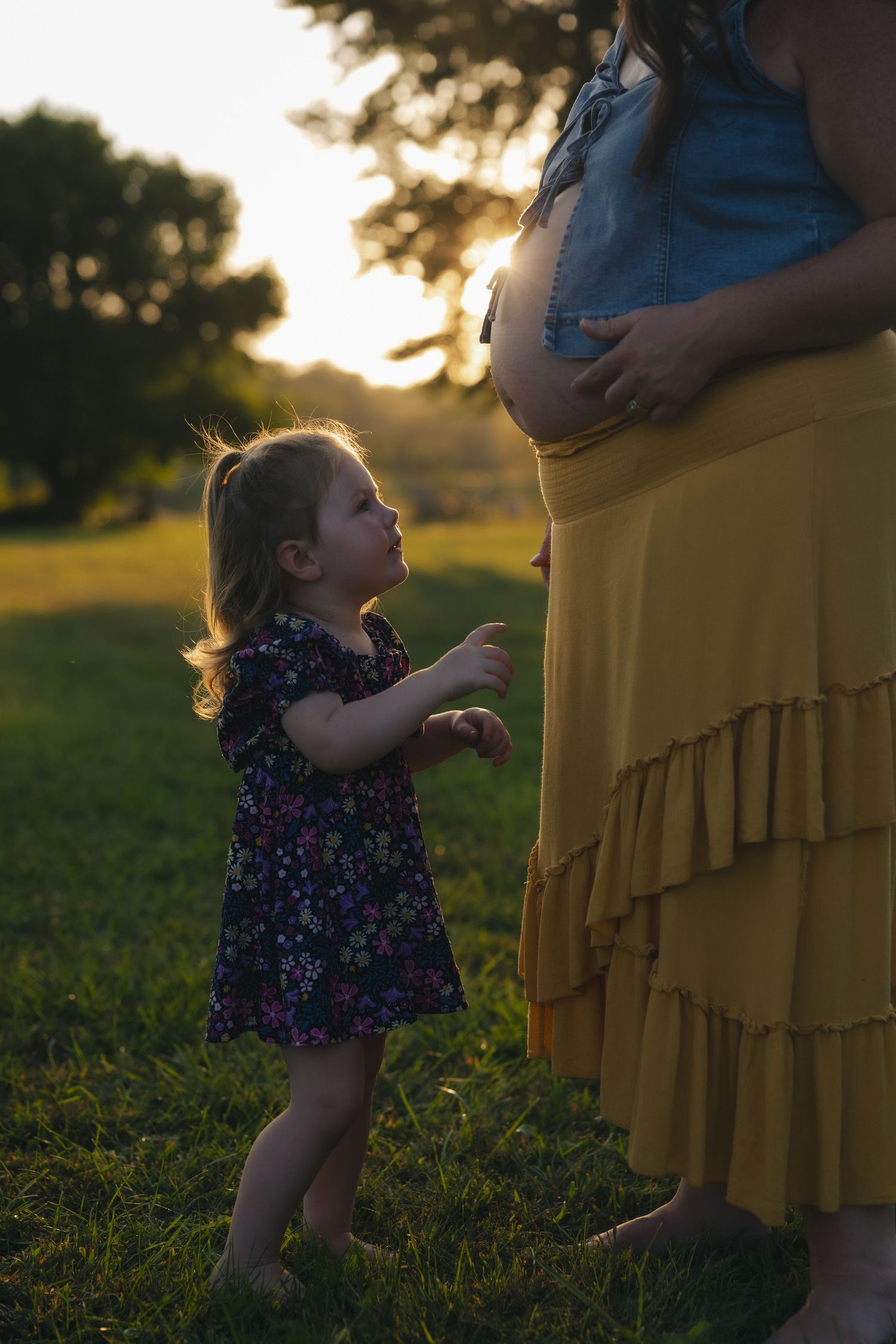 A toddler in a floral dress explores while backlit by warm golden sunlight in a grassy field at sunset.