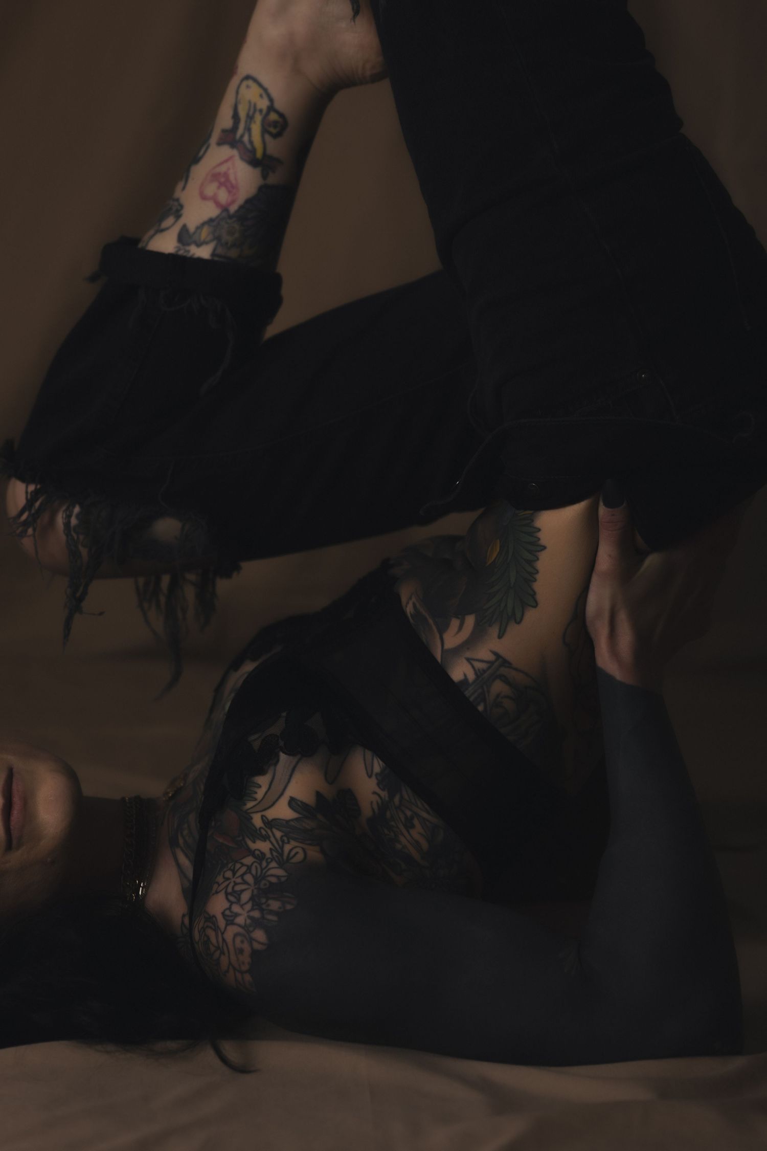 Person lying down with extensive floral tattoos on torso and arm, wearing black frayed flare pants in moody lighting.