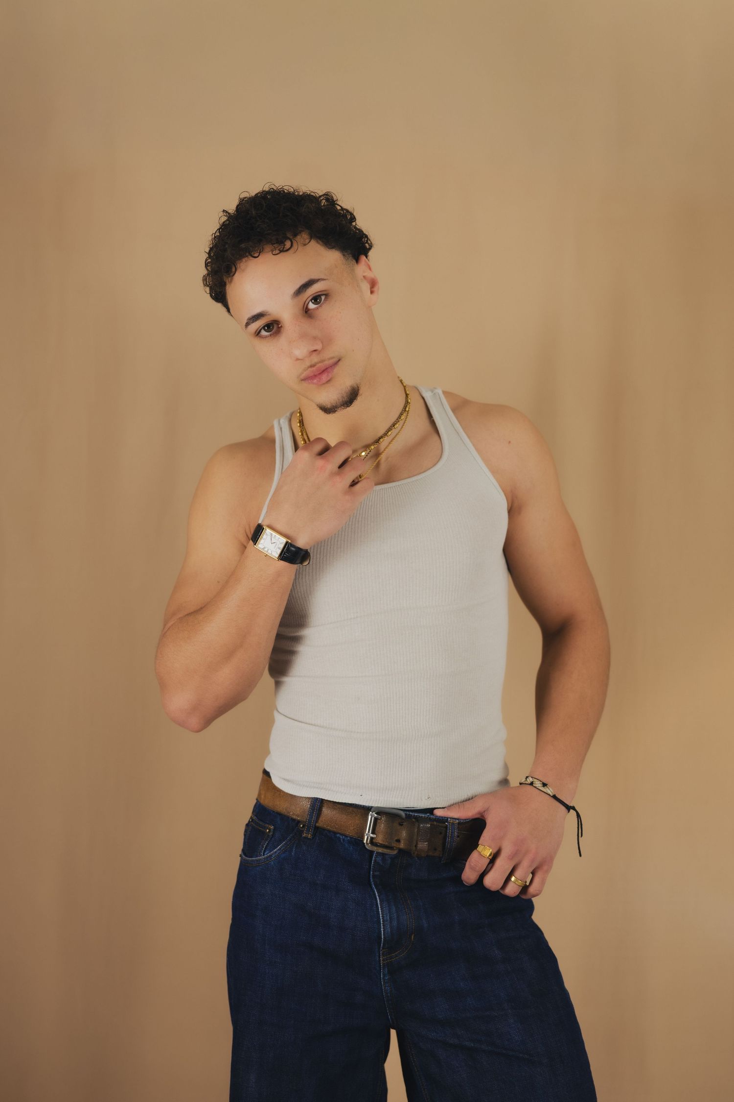 Young man in white tank top and jeans posing with gold chain necklace and watch against beige background.