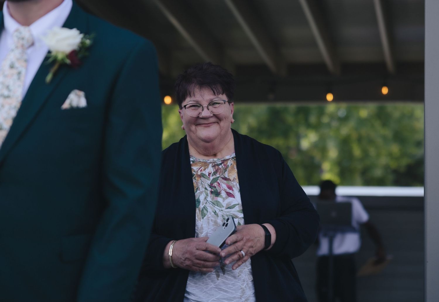 Smiling older woman in floral top at outdoor wedding venue with man in dark green suit in foreground.