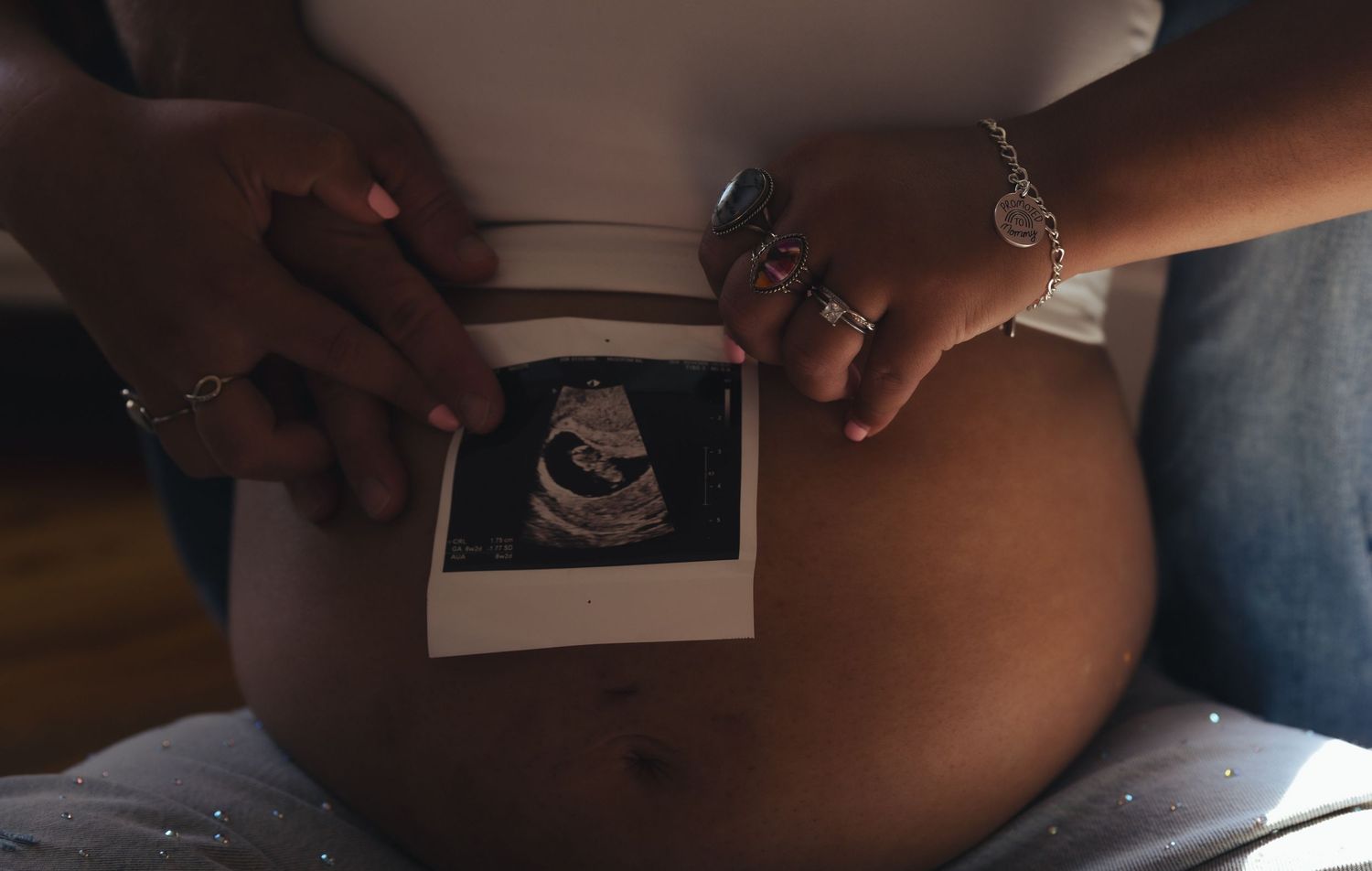 Pregnant woman holding ultrasound sonogram photo against bare baby bump, two hands with jewelry visible.