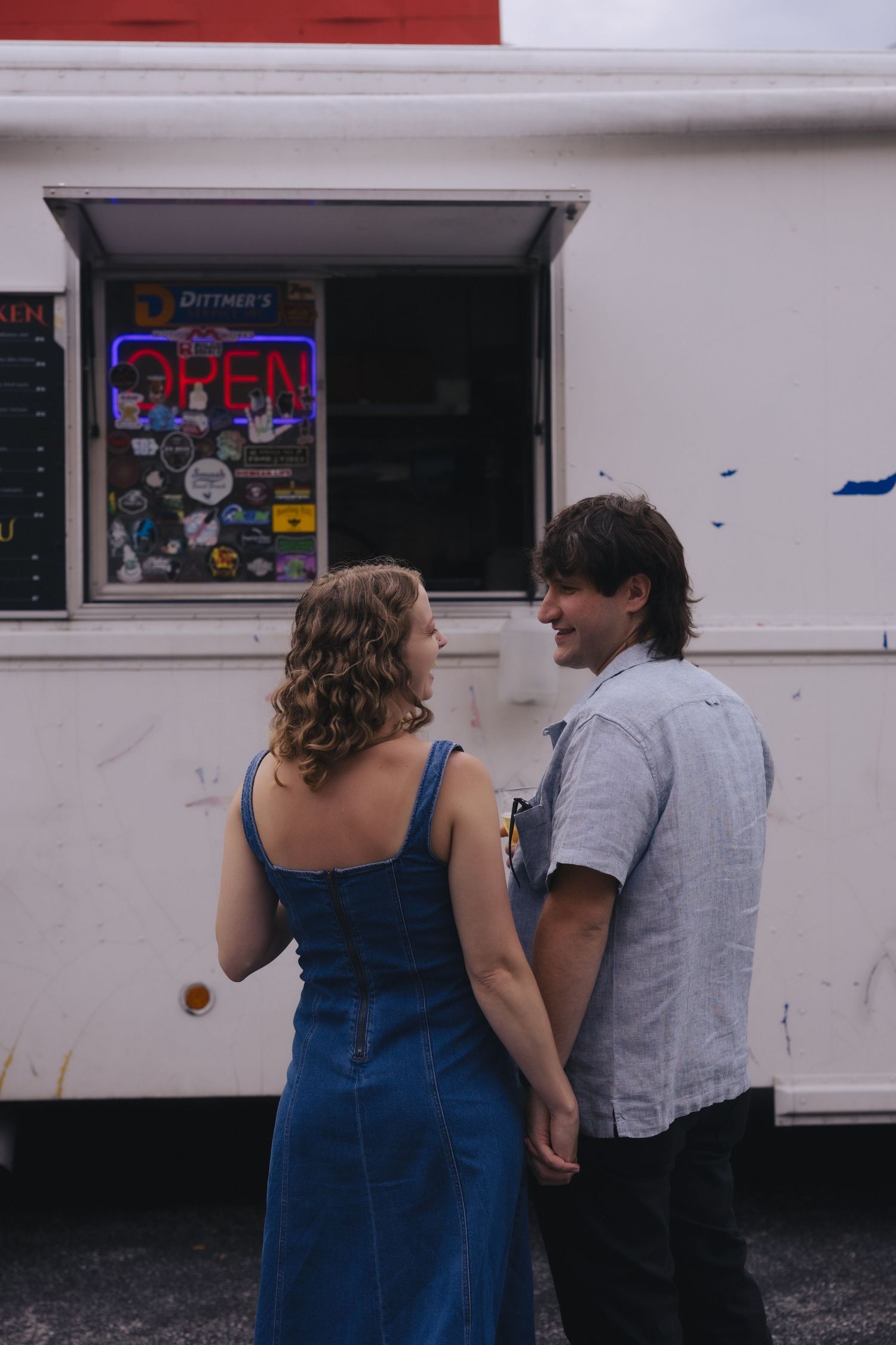 Couple holding hands and smiling in front of an open food truck with a neon open sign.