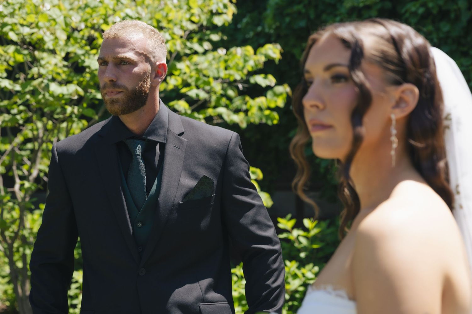 A groom in a dark suit gazes at his bride in a white wedding gown outdoors surrounded by lush green trees.