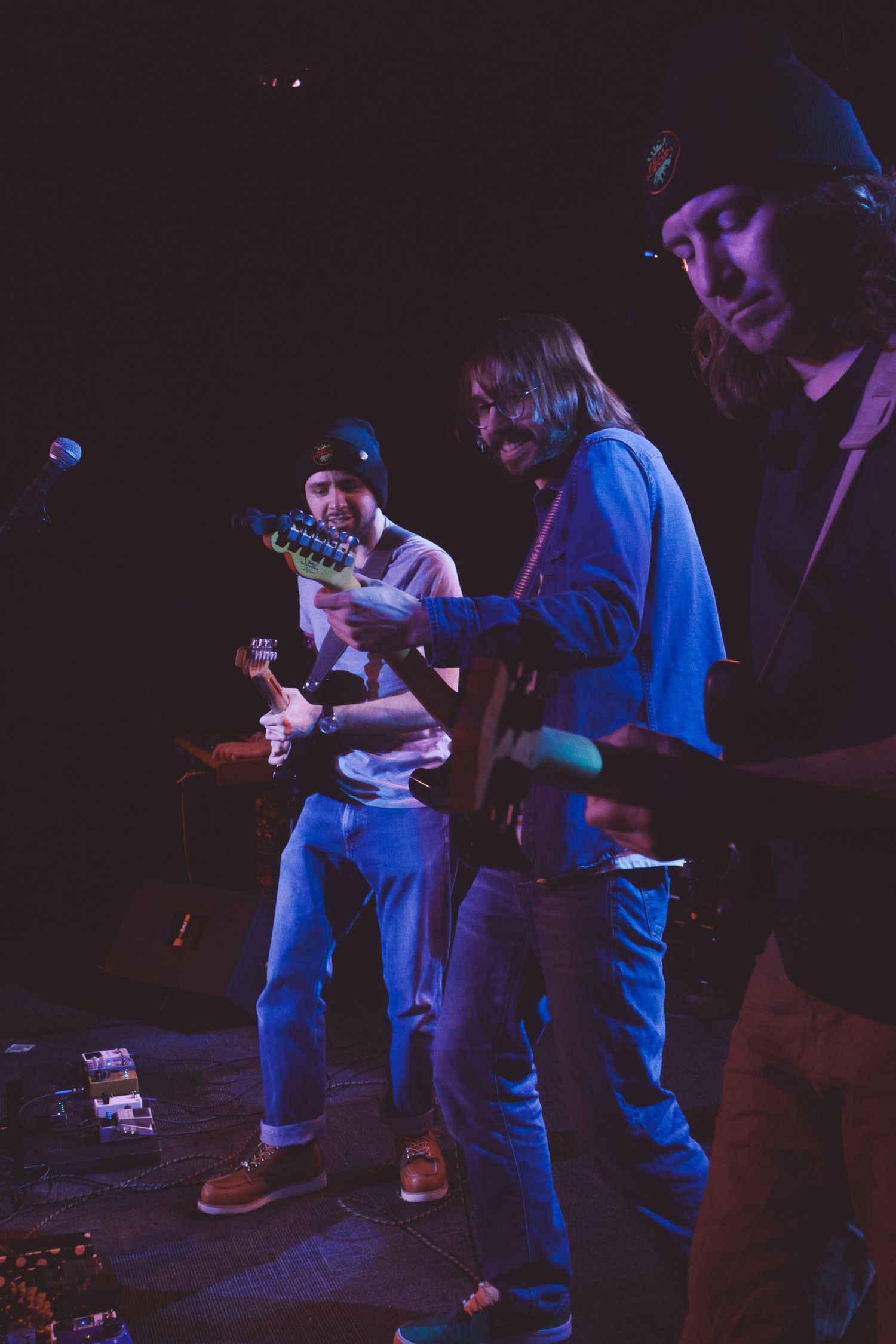 Musicians performing on stage in moody blue lighting, with guitarists in casual attire during a live concert performance.