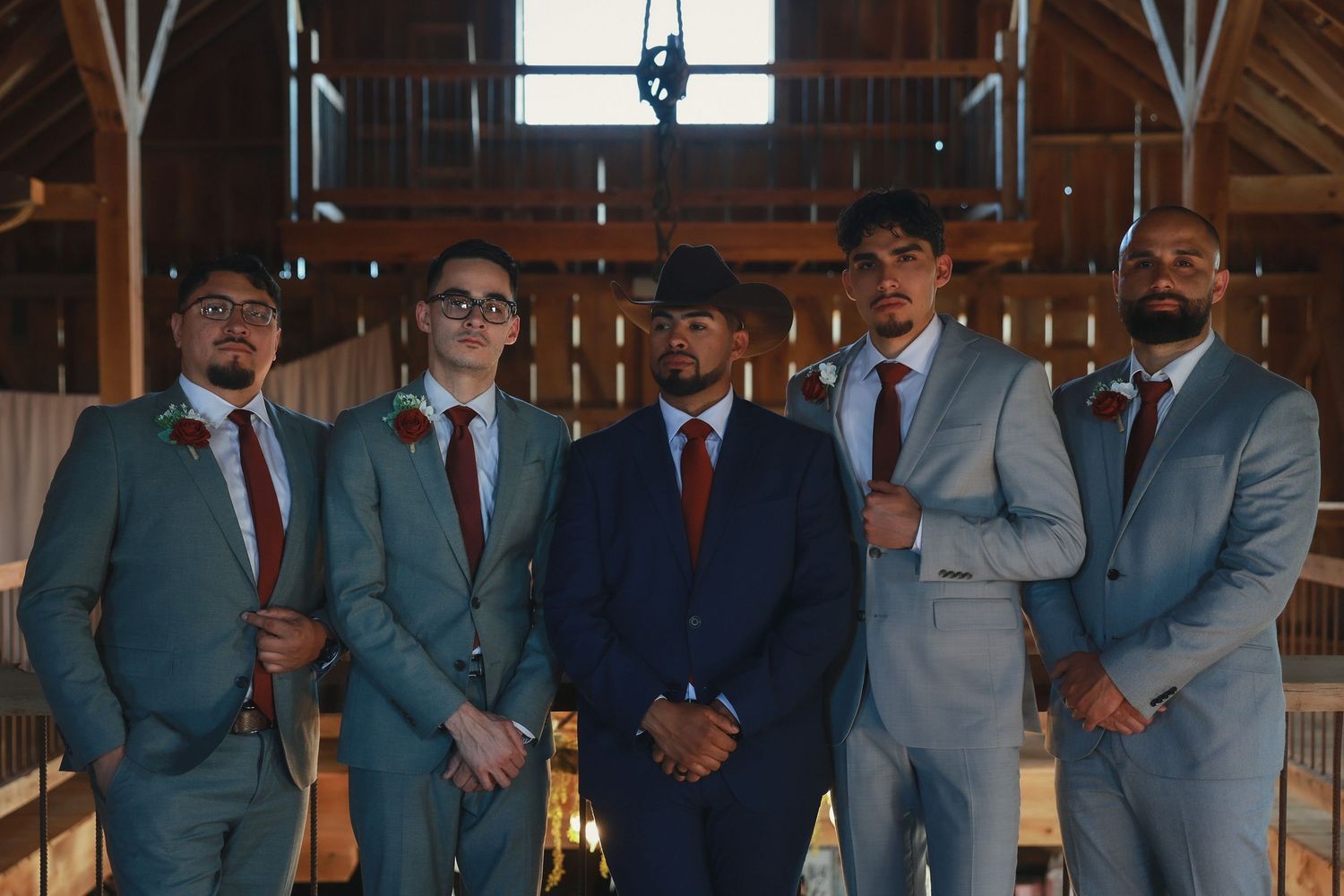 A group of five groomsmen pose together in coordinated suits with red ties inside a rustic wooden barn venue.