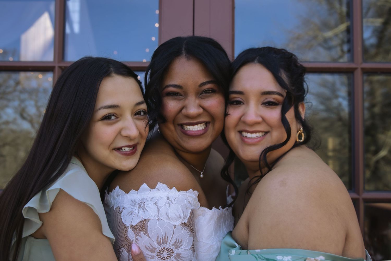 Three friends share joyful smiles while posing close together in formal attire at an outdoor venue.