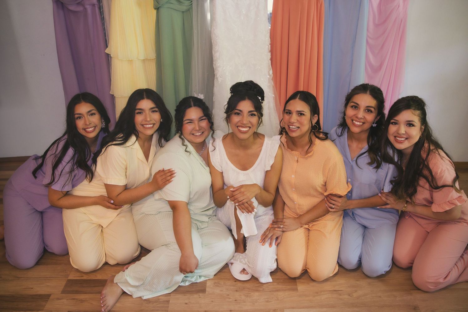 A group of seven friends wearing coordinated pastel colored loungewear sit together smiling on a wooden floor.