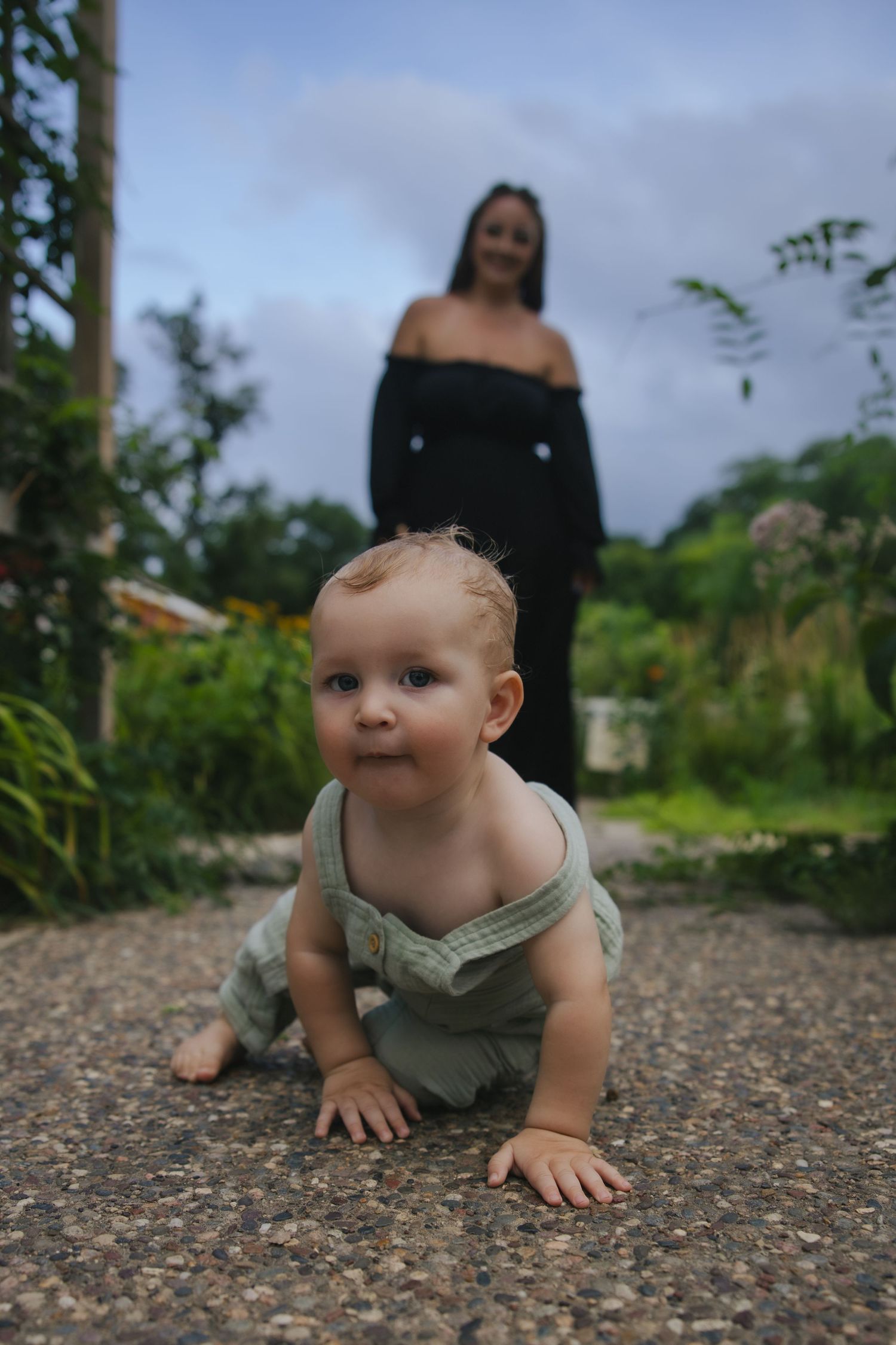 Baby in a gray outfit crawls on a gravel path while exploring outdoors on a cloudy day.