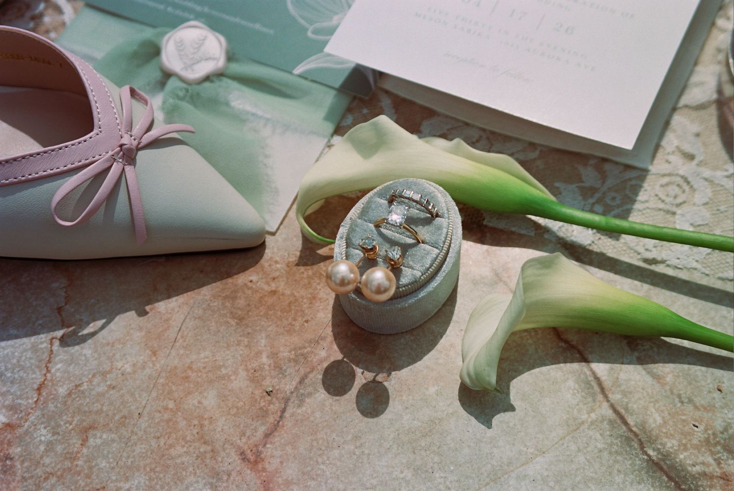Wedding rings and pearl earrings in a ring dish beside calla lilies, pink heels, and elegant stationery on stone.