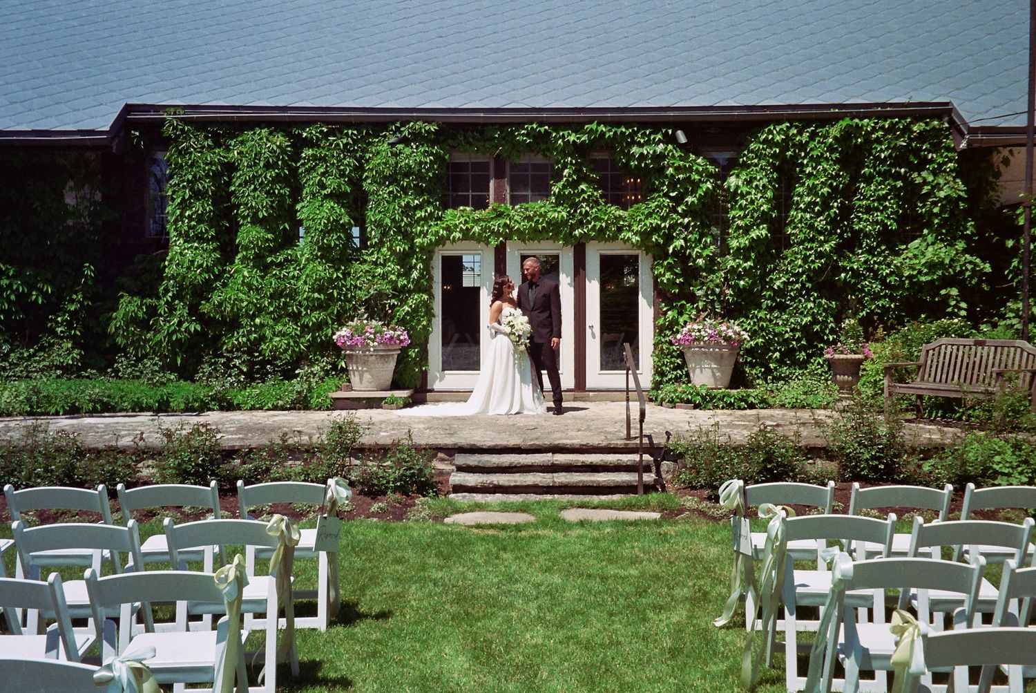Bride and groom pose on ivy-covered venue steps surrounded by white chairs set for an outdoor garden wedding ceremony.