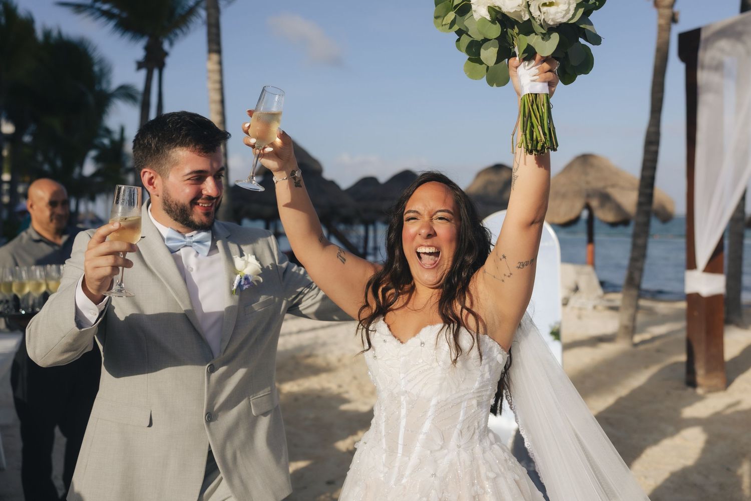 Joyful newlyweds celebrate on a tropical beach, bride raising bouquet and groom toasting with champagne at their destination wedding.