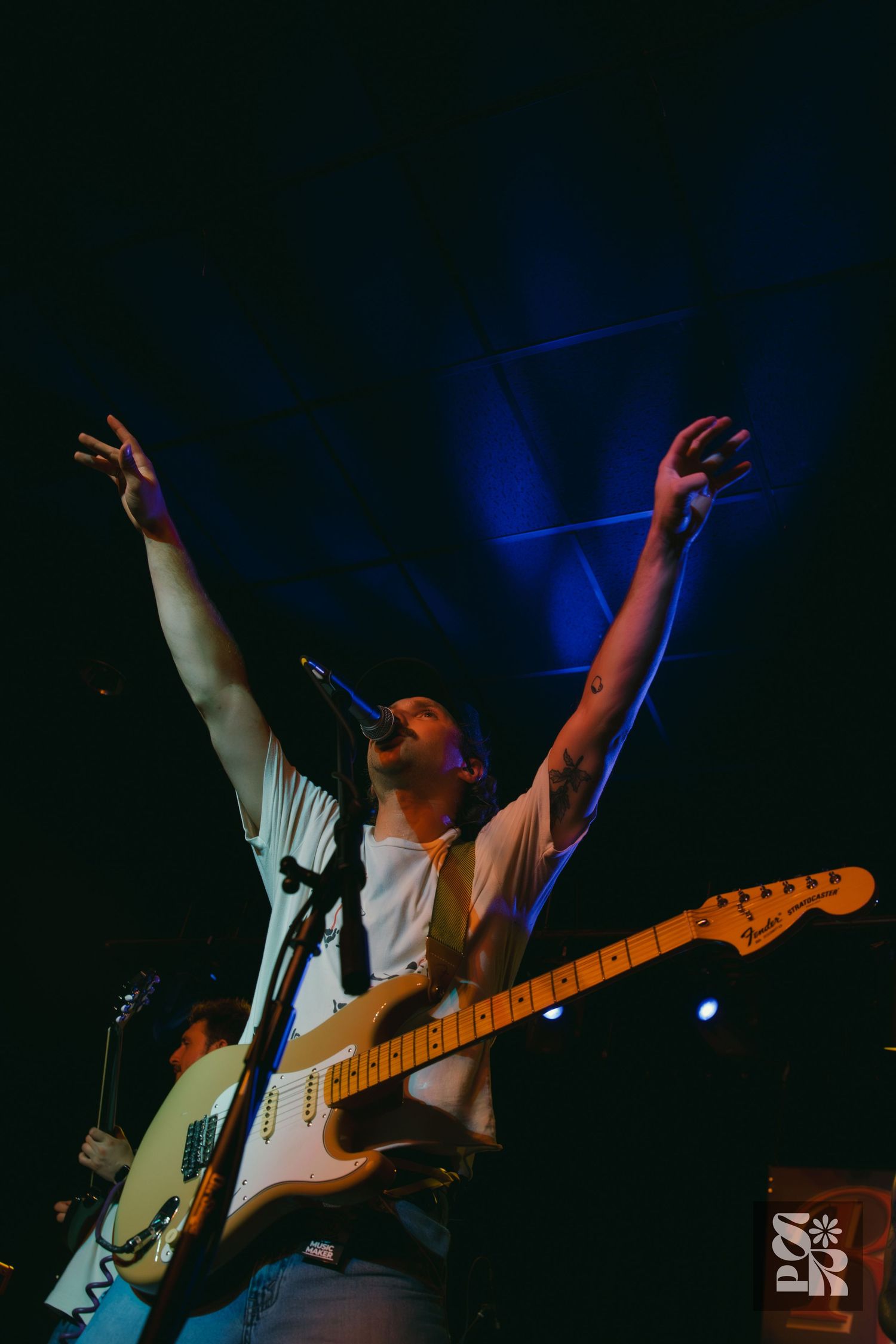 A passionate musician plays electric guitar and sings while reaching up with raised arms under dramatic blue stage lighting.
