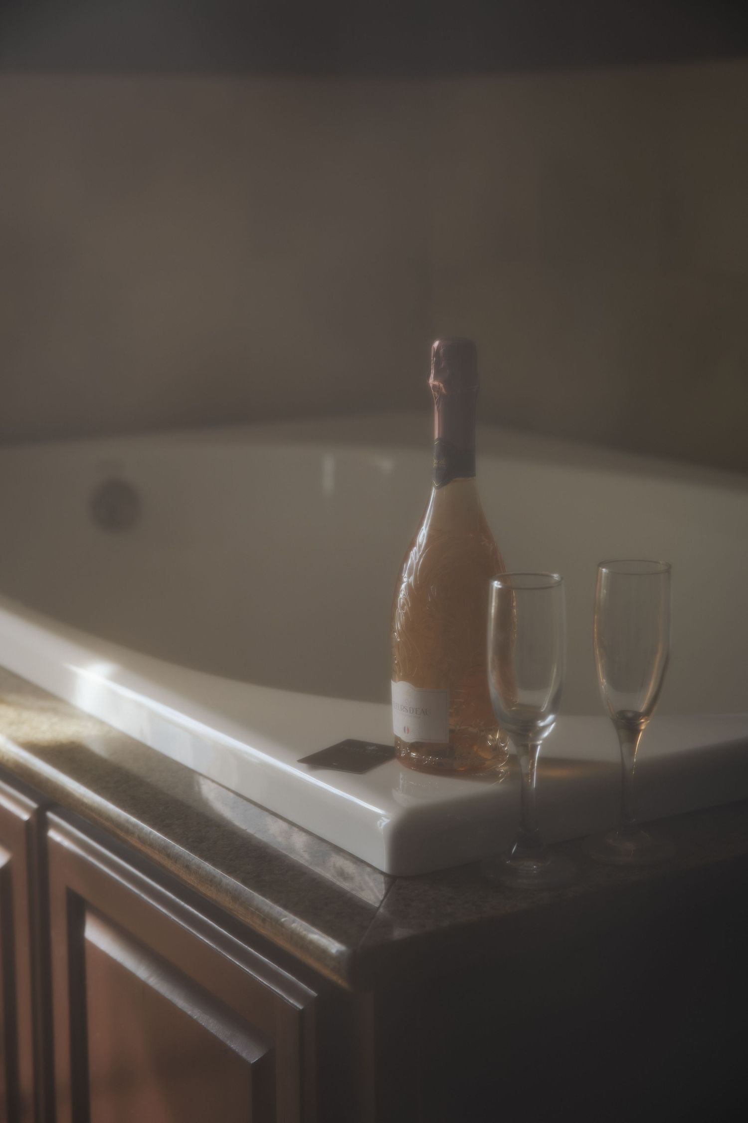 A bottle of rose wine and two empty champagne flutes sit atop a dark wooden surface in moody lighting.