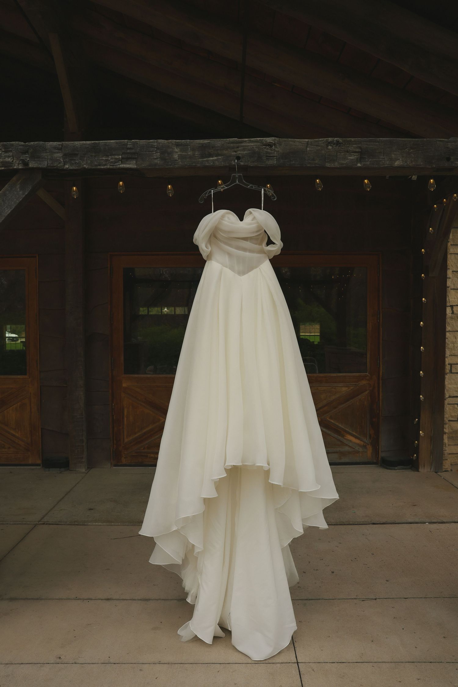Elegant white wedding dress hanging from rafters on display at a rustic wooden venue with twinkling lights.