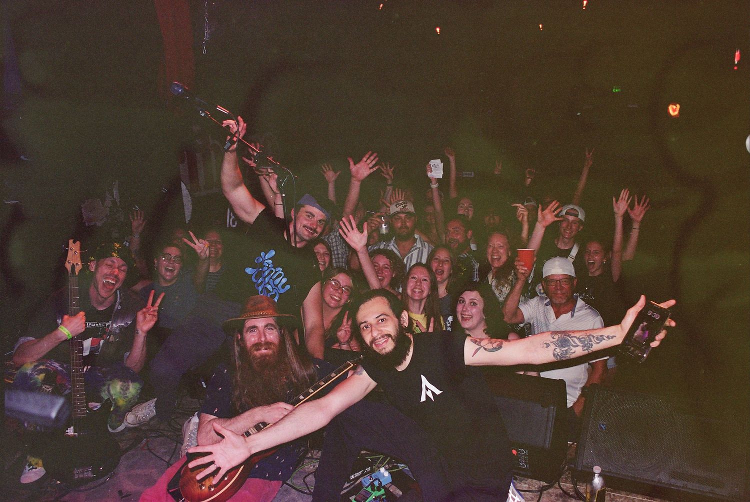 Excited crowd at a live music concert raising hands, smiling and celebrating together in a dimly lit venue.