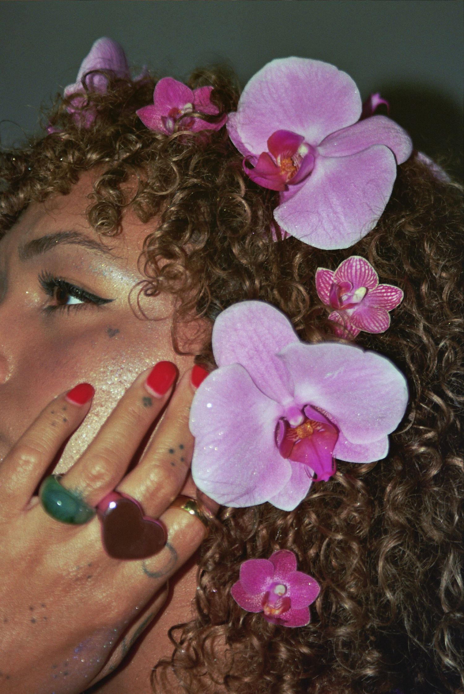 Woman with curly hair adorned with pink orchid flowers, red nails, and colorful rings covering her face.