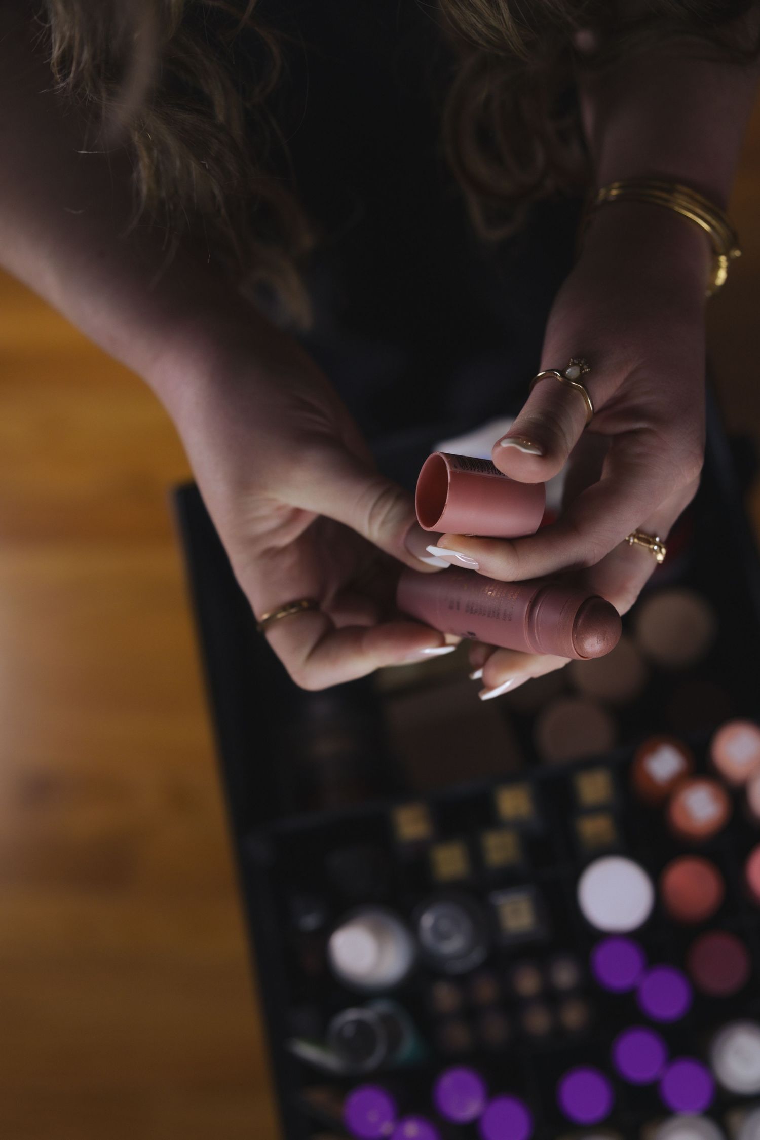 Close-up of woman's hands holding lipstick tubes over a colorful makeup palette with gold rings and bracelet.