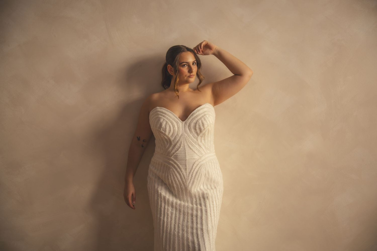 A bride poses in a fitted strapless white gown with geometric patterns against a beige textured wall.