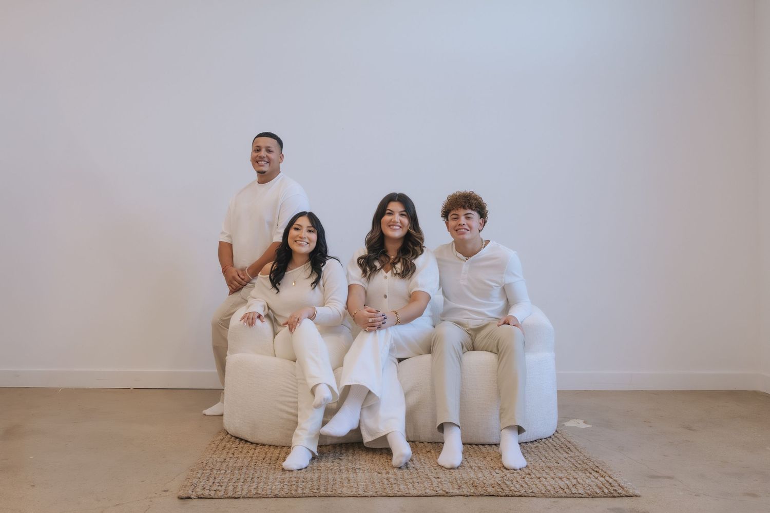 Four friends in white outfits pose together on neutral furniture against a white wall for a stylish group photo.