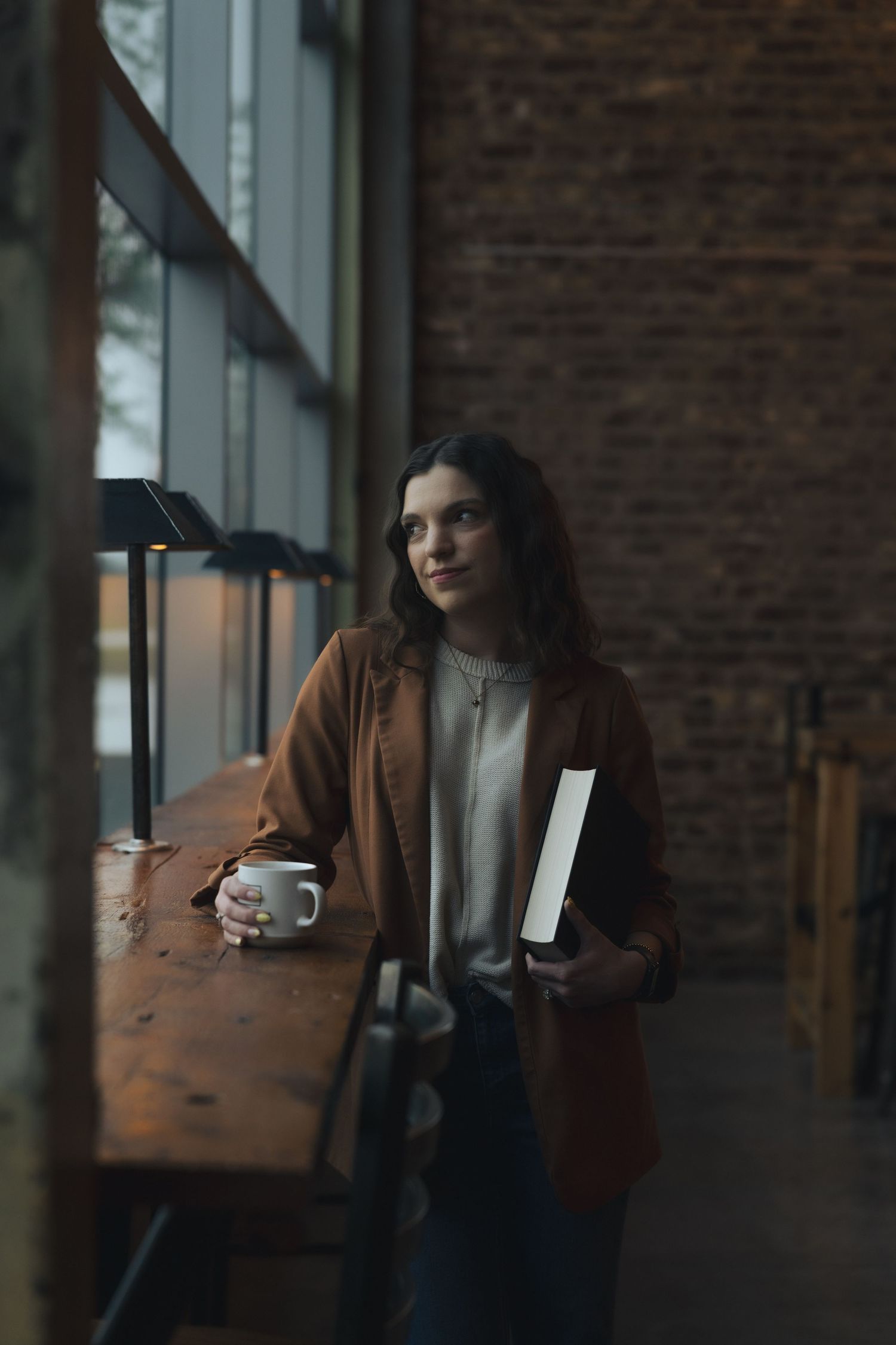 Young woman in brown blazer holds coffee mug and book in a cozy cafe with brick walls and wooden counter.