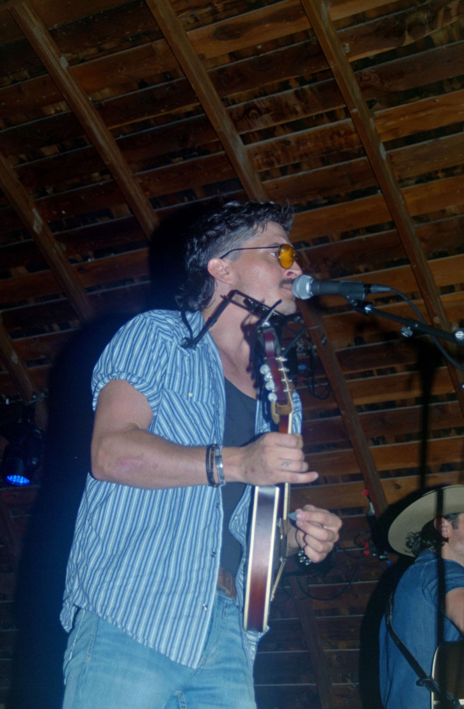 Male guitarist with sunglasses performs on stage in a rustic wood-beamed venue, singing into a microphone while playing acoustic guitar.