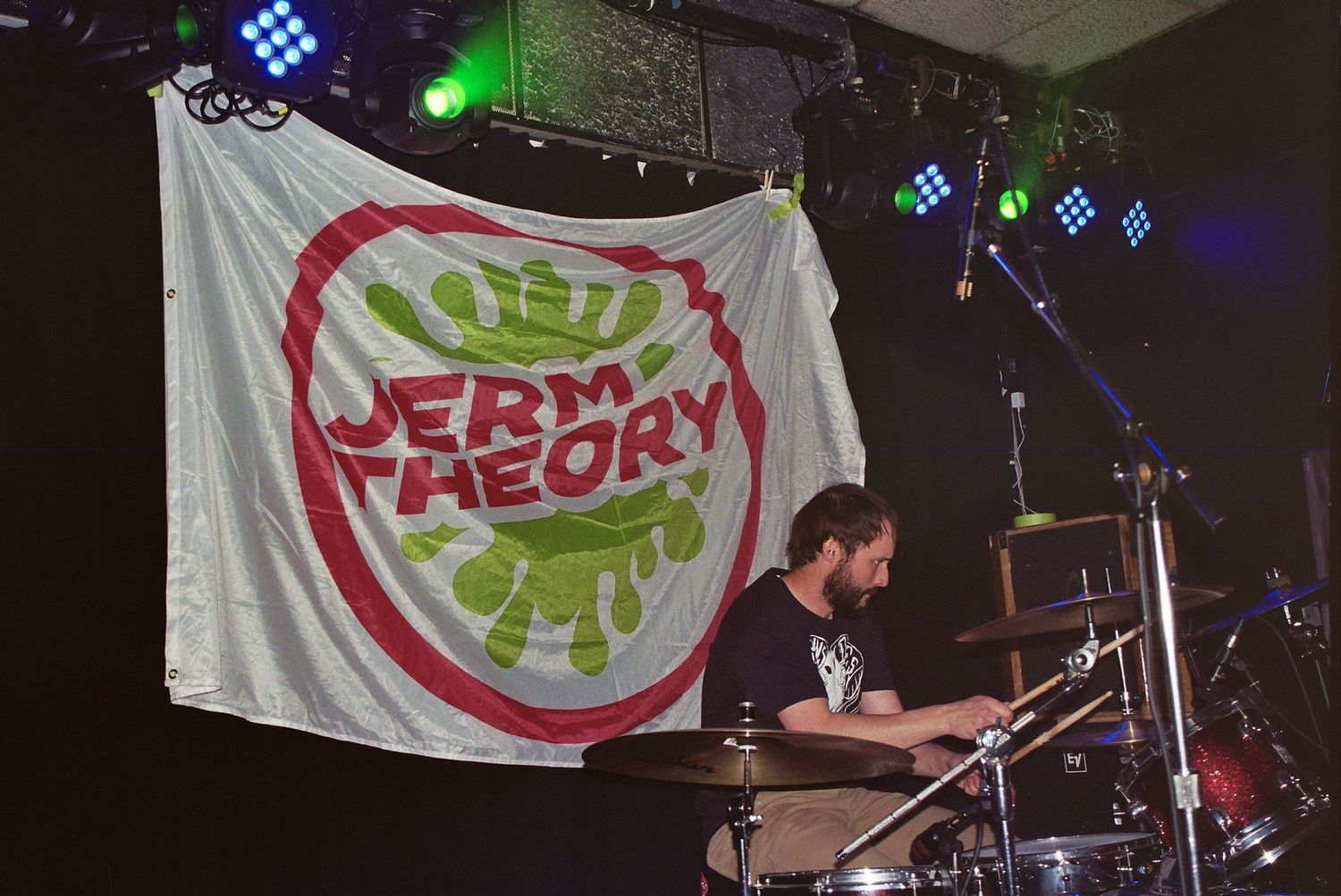 Drummer performing on stage behind a Jerm Theory band logo flag at a live music venue.