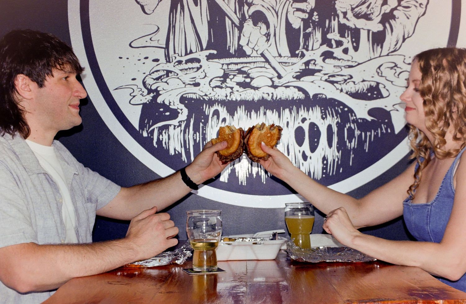 A couple toasting burgers at a restaurant table with beer glasses, smiling in front of a decorative mural.