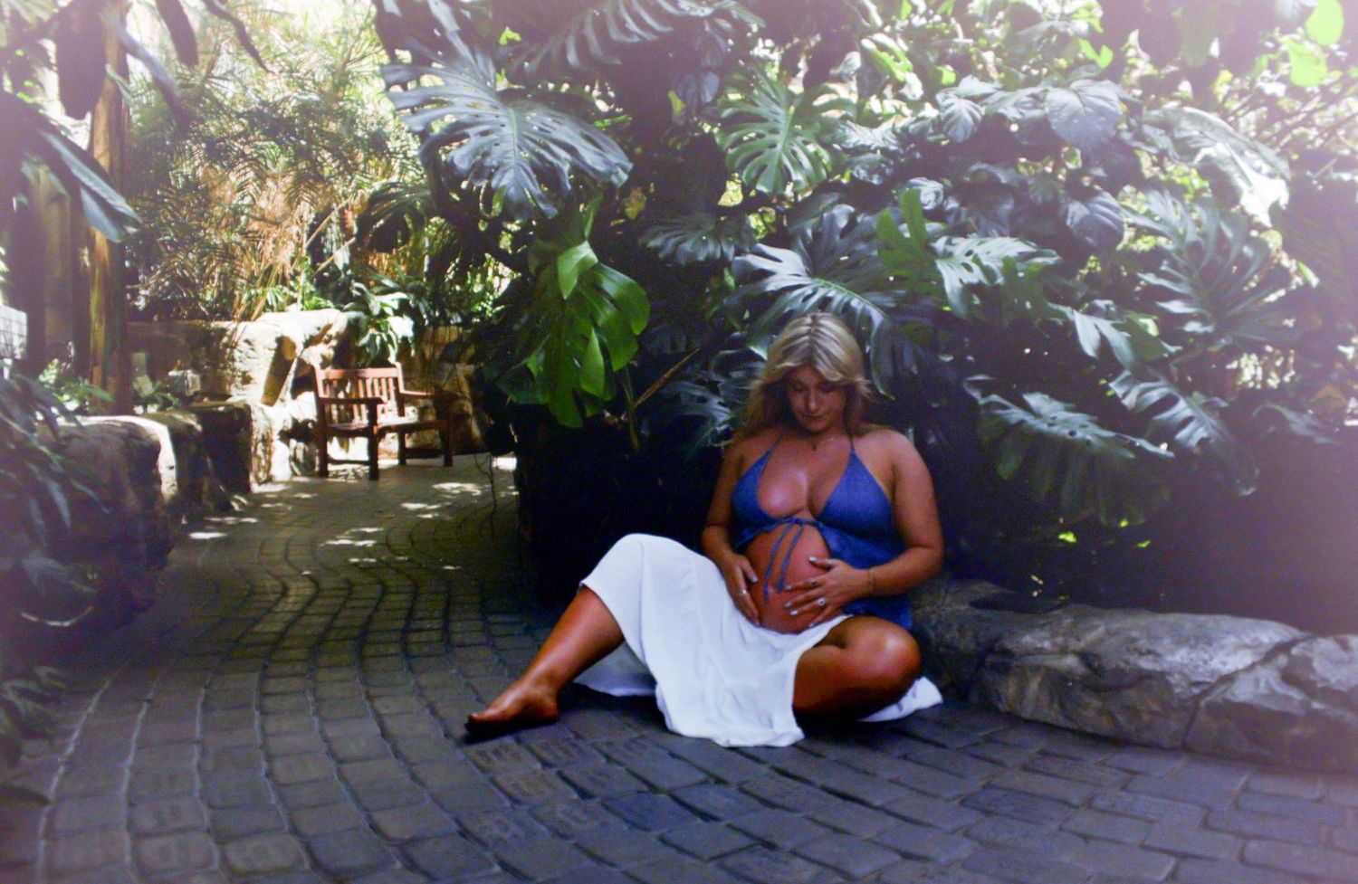 Pregnant woman in blue top and white skirt sitting on brick path surrounded by tropical green plants.