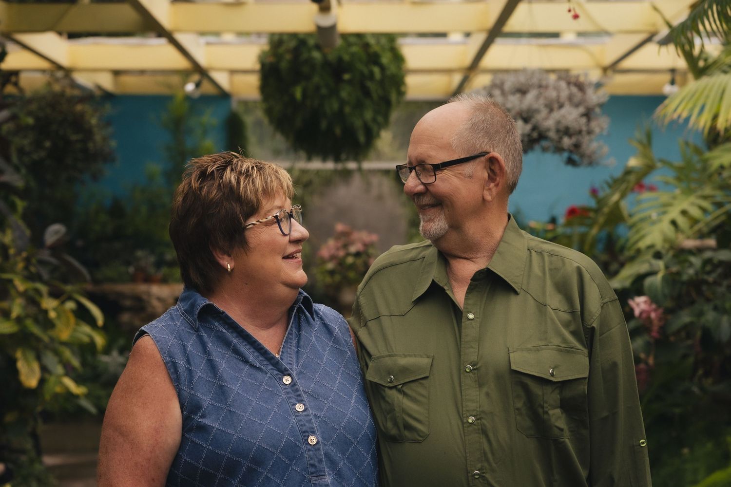 Smiling older couple standing together in a lush greenhouse surrounded by green plants and flowers.