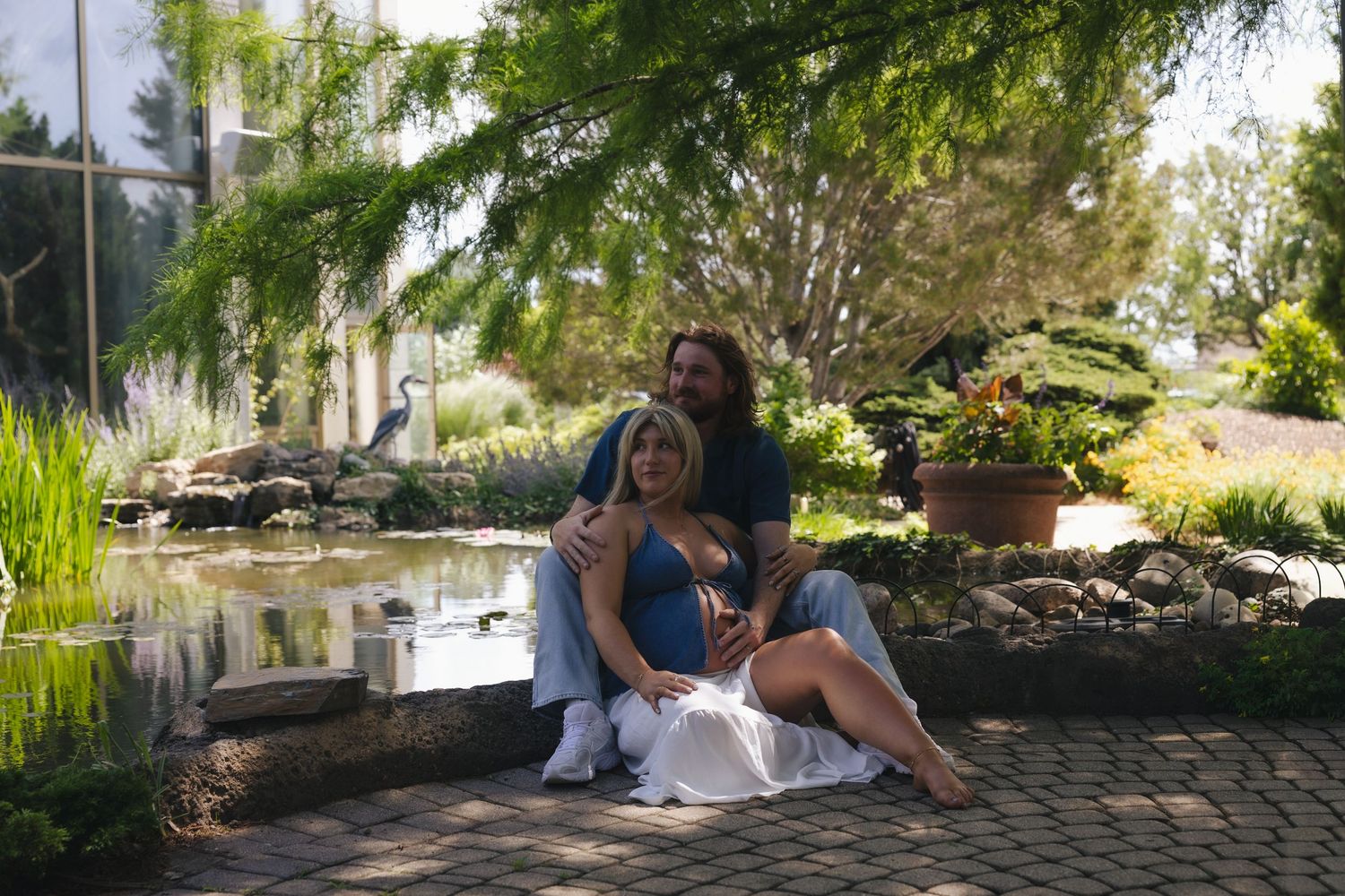 Couple sitting together by a garden pond surrounded by lush greenery, woman in blue dress during maternity photoshoot.