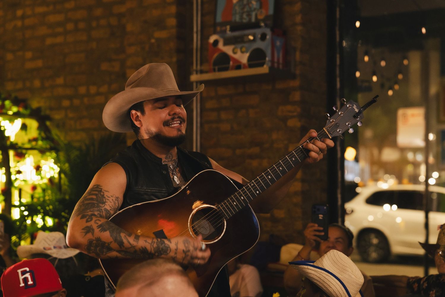 Tattooed male musician in cowboy hat playing acoustic guitar at an outdoor venue at night.